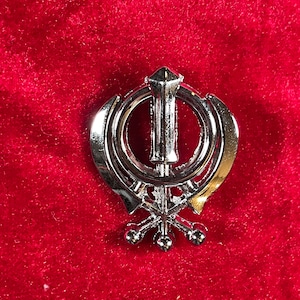 Puede incluir: Un Khanda de plata, símbolo del sijismo, sobre un fondo de terciopelo rojo.