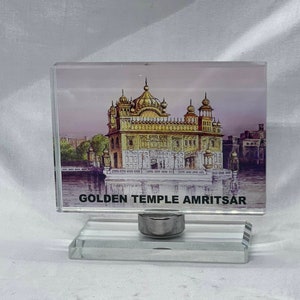 Può includere: Un fermacarte di vetro con un'immagine del Tempio d'Oro ad Amritsar, in India. Il tempio è dorato e ha una grande cupola. Il fermacarte è su una base di vetro trasparente.