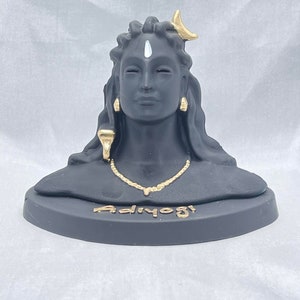 May include: A black statue of Adiyogi, a bust of a deity with closed eyes, adorned with gold-colored jewelry and a crescent moon. The base of the statue has the word "Adiyogi" in gold lettering.