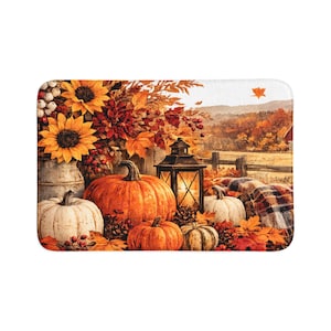 Op de afbeelding: Een rechthoekige badmat met een herfsttafereel met gele zonnebloemen, oranje en witte pompoenen, een zwarte metalen lantaarn en een plaid. De achtergrond toont een landelijk landschap met herfstbladeren en een houten hek.