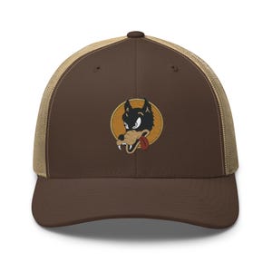 Könnte beinhalten: Eine braune Trucker-Cap mit beigem Netzrücken und brauner Vorderseite. Mittig auf der Vorderseite befindet sich ein gestickter, runder Aufnäher, der einen stilisierten schwarz-beigen Wolfskopf mit roter Zunge und weißen Fangzähnen zeigt.