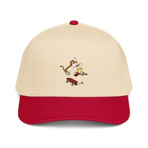 Puede incluir: Gorra de béisbol con corona color crema y visera roja. La gorra presenta una ilustración de dibujos animados de un niño y un tigre jugando con un carro rojo. La gorra tiene un botón rojo en la parte superior.