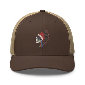 May include: A brown trucker hat with a tan mesh back and an embroidered graphic on the front. The embroidery depicts a skull in profile wearing a traditional-style headdress with red, white, blue, and green feathers, centered on the front panel.
