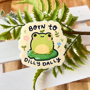 May include: A sticker with a cartoon frog on a lily pad, with the words "Born to Dilly Dally." The frog is green with a yellow belly, and the sticker has a light yellow background with floral accents. The sticker is surrounded by green foliage.