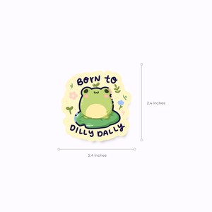 May include: A die-cut sticker featuring a cartoon frog on a lily pad. The frog is green with a cream-colored belly. The sticker is 6.1 cm in diameter and has the text "BORN TO DILLY DALLY".