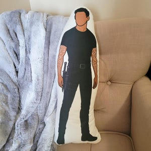 May include: A decorative pillow featuring a stylized illustration of a person in black clothing with a sword. The pillow has a white background and the person has tattoos on their arms. The pillow is resting on a beige chair.
