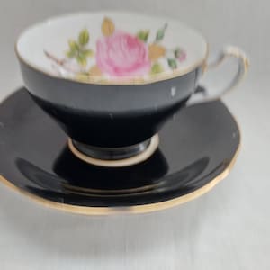 May include: Black teacup and saucer set with gold trim. The teacup interior features a floral design with a pink rose. The saucer has a matching gold trim. A classic and elegant tea set.