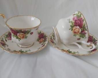 May include: Two white teacups and saucers with a floral rose pattern in shades of red, yellow, and green. The rims and handles are trimmed with gold. One cup is upright, the other is tilted on its side.