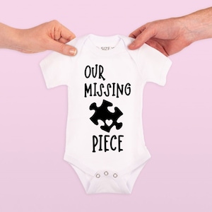 May include: White baby bodysuit featuring the text "OUR MISSING PIECE" and a black puzzle piece graphic with a heart. The bodysuit is held up by two hands against a light pink background.