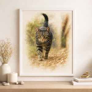 May include: Watercolor print of a tabby cat walking forward, framed in a white border. The cat has brown and black stripes with yellow eyes. The background is a soft, blurred depiction of a path and foliage, with a warm, neutral color palette.