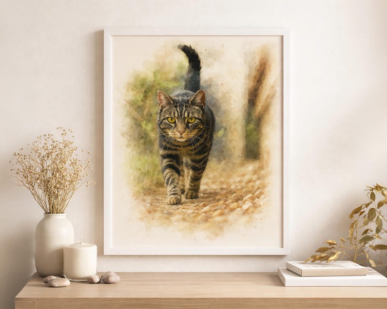May include: Watercolor print of a tabby cat walking forward, framed in a white border. The cat has brown and black stripes with yellow eyes. The background is a soft, blurred depiction of a path and foliage, with a warm, neutral color palette.