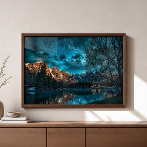 May include: A framed landscape print depicts a mountain range reflected in a lake under a starry night sky. The artwork features a dark brown frame and is displayed on a wooden cabinet. The color palette includes blues, greens, and browns.