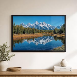 May include: A framed landscape photograph featuring snow-capped mountains reflected in a calm lake. The image showcases a vibrant blue sky, lush green trees, and golden-brown foliage, all enclosed in a black frame, creating a serene nature scene.