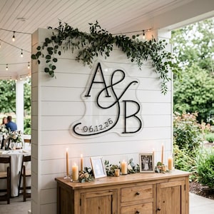 May include: A clear acrylic sign featuring the black lettering A & B and the date 06.12.26, mounted on a white shiplap wall decorated with green eucalyptus branches. The sign is positioned above a wooden cabinet with candles and framed photos.
