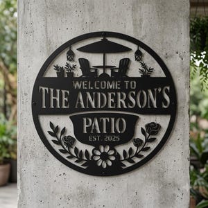 Può includere: Un'insegna da parete circolare in metallo nero con un design intagliato raffigurante due sedie, un tavolo e un ombrellone. Il testo recita "WELCOME TO THE ANDERSON'S PATIO EST. 2025" con decorazioni floreali di rose e foglie sul fondo.