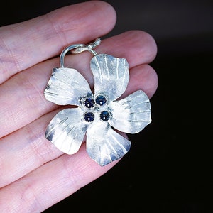 May include: A silver flower pendant with four dark blue gemstones in the center. The flower has five petals and a textured surface.