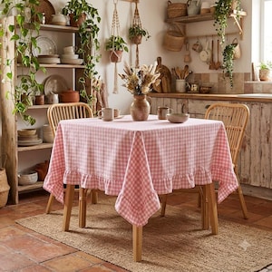 Puede incluir: Un mantel cuadrado con un patrón de cuadros vichy en rosa claro y blanco con un borde con volantes. La tela está colocada sobre una mesa de madera de cuatro patas, situada entre dos sillas de madera en una cocina.