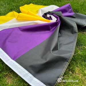 May include: A purple, black and white pride flag with yellow trim. The flag is folded and lying on a bed of green grass.