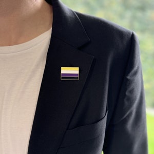 May include: A rectangular enamel pin displaying the non-binary pride flag. The pin has a black border and features horizontal stripes of yellow, white, purple, and black. The pin is attached to a dark navy blazer.