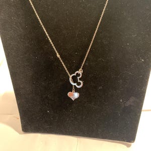 May include: A silver-colored necklace featuring a Mickey Mouse head pendant with a heart-shaped charm. The necklace is displayed on a black velvet surface, showcasing its delicate design and shine. The chain is thin and elegant.
