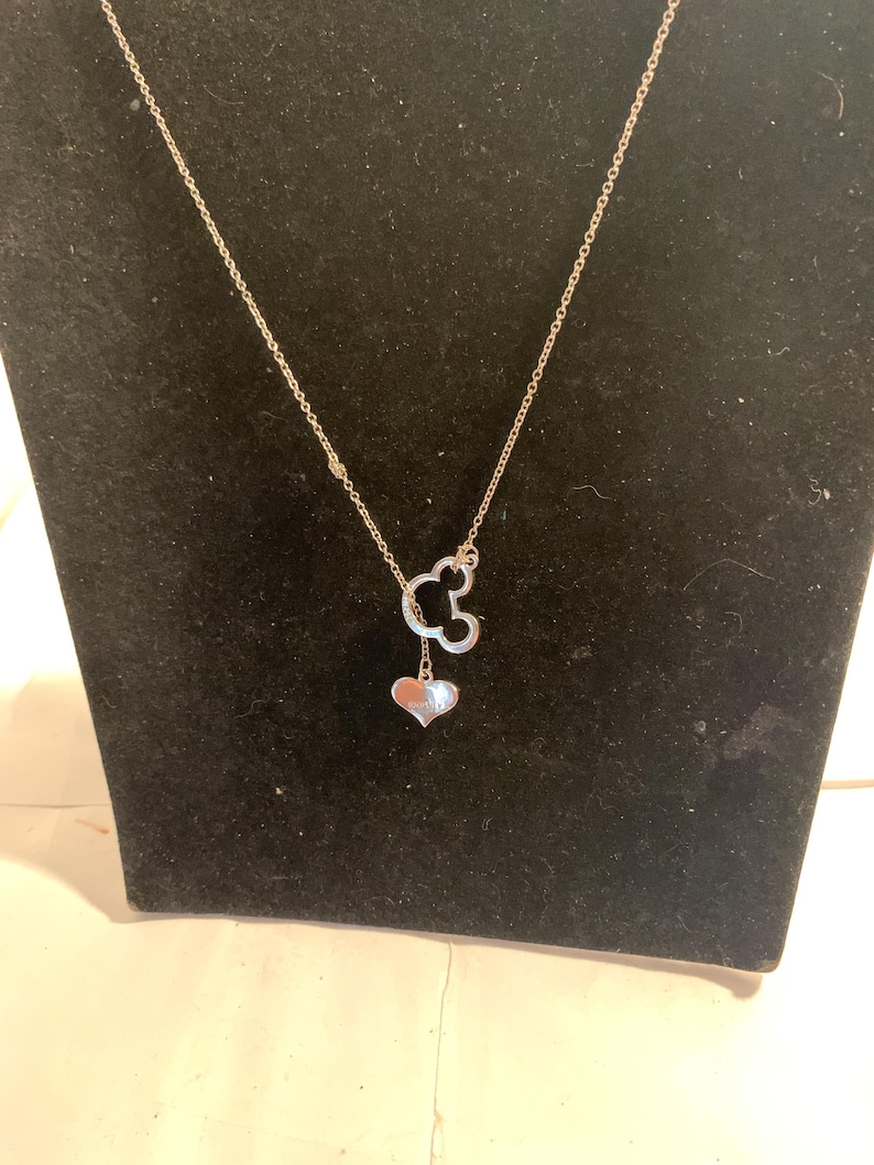 May include: A silver-colored necklace featuring a Mickey Mouse head pendant with a heart-shaped charm. The necklace is displayed on a black velvet surface, showcasing its delicate design and shine. The chain is thin and elegant.