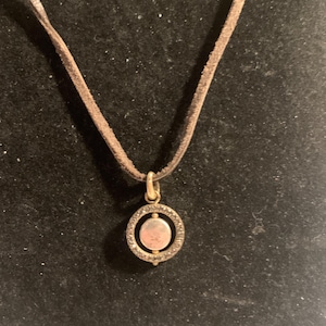 May include: A pendant necklace featuring a circular pendant with a central, light-colored stone surrounded by a ring of smaller stones. The pendant hangs from a brown suede cord. The metal components appear to be gold-toned.