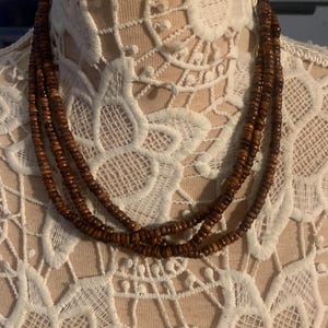 May include: A three-strand necklace made of small, round, dark brown wooden beads. The necklace is displayed on a white lace surface, showcasing the natural wood tones and the intricate design of the jewelry.