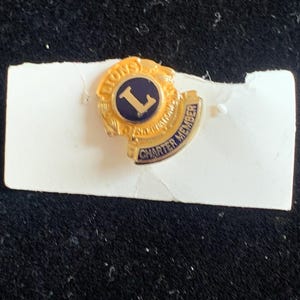 May include: A gold-tone metal lapel pin featuring the Lions International logo with a dark blue circular centre, a gold letter L, and a curved banner at the bottom with the text CHARTER MEMBER in blue lettering against a gold background.