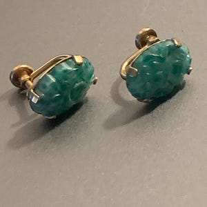 May include: A pair of vintage earrings featuring carved green gemstones set in gold-tone metal. The gemstones have an oval shape and are intricately detailed. The earrings have a screw-back closure.