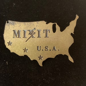 Könnte beinhalten: Ein goldfarbener Metallausschnitt der Vereinigten Staaten mit der Aufschrift "MIXIT" und "U.S.A.".