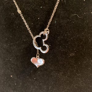 May include: A silver necklace featuring a Mickey Mouse silhouette pendant with a dangling heart charm. The heart charm is engraved with the word "Disney". The necklace is displayed against a black background.