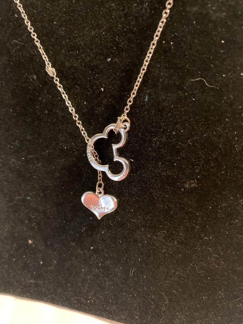 May include: A silver necklace featuring a Mickey Mouse silhouette pendant with a dangling heart charm. The heart charm is engraved with the word "Disney". The necklace is displayed against a black background.