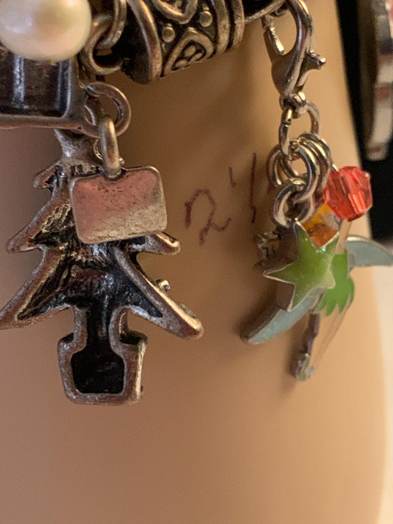 Puede incluir: Primer plano de una pulsera de plata con varios abalorios. Los abalorios incluyen un &aacute;rbol de Navidad, una estrella con detalles verdes y naranjas, y un peque&ntilde;o abalorio rectangular. La pulsera tambi&eacute;n presenta una perla y elementos met&aacute;licos decorativos.