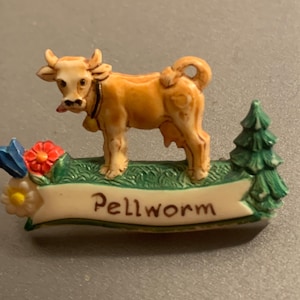 Könnte beinhalten: Beigefarbene Kuhfigur mit Glocke um den Hals, steht auf einer grünen Basis mit Banner "Pellworm". Die Basis hat eine rote Blume, einen blauen Schmetterling und einen grünen Baum.