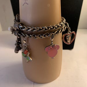 Puede incluir: Una pulsera de dijes plateada con m&uacute;ltiples hilos y dijes decorativos. Los dijes incluyen una cabeza de Minnie Mouse rosa, una flor roja y verde y un dije en forma de coraz&oacute;n. La pulsera se muestra en un brazo de maniqu&iacute; beige.