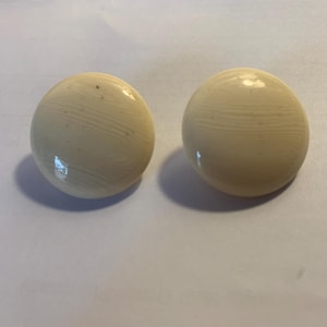 Puede incluir: Dos objetos redondos de color crema con un acabado brillante y un sutil patrón similar a la veta de la madera. Los objetos son lisos y parecen estar hechos de un material sólido. Se encuentran sobre un fondo liso blanquecino.