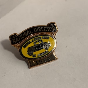 May include: A gold-toned pin with a banner reading "NATIONAL DIRECTOR." The pin features a yellow oval with a black Model A Ford car and the text "MODEL A FORD CLUB OF AMERICA 1979."