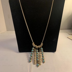 Puede incluir: Un collar de cadena dorada con un colgante de piedra turquesa. El colgante presenta múltiples piedras turquesas y barras doradas que cuelgan de un anillo de cuentas doradas.