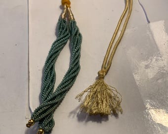 May include: A decorative tassel with a braided teal cord and a gold-colored tassel. The cord is braided into a thick rope and has gold-colored end caps. The tassel is made of a gold-colored thread.