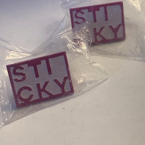 May include: Two red and white enamel pins with the word "STICKY" in white letters on a red background.
