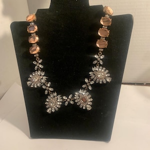 May include: A statement necklace with a black display stand. The necklace features large, oval-shaped peach-colored stones and clusters of clear, sparkling crystals. The crystals are arranged in floral patterns, creating a glamorous and eye-catching design.