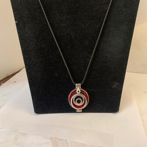 Könnte beinhalten: Eine silberne Halskette mit einem roten und schwarzen runden Anhänger. Der Anhänger hat konzentrische Kreise mit silbernem Rand.