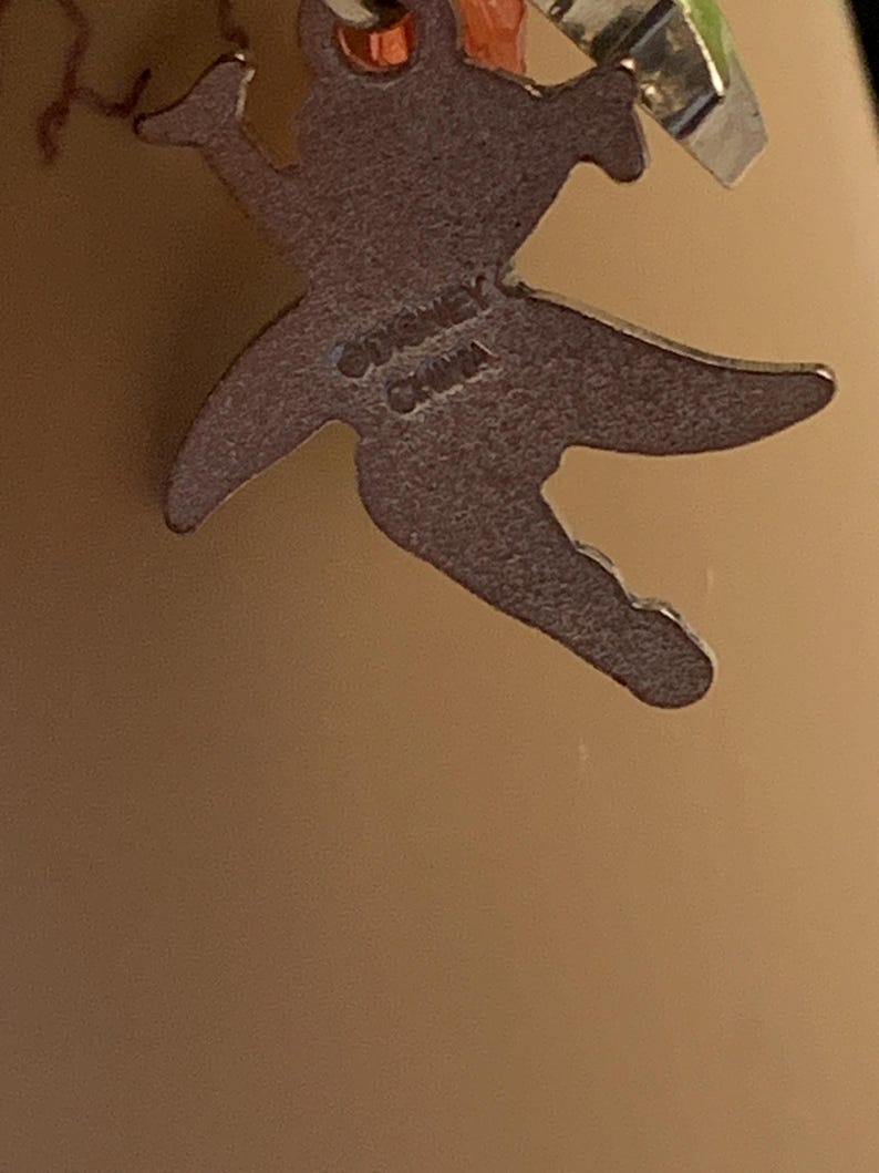 Puede incluir: Primer plano de un dije plateado con forma de hada. El dije tiene el texto "&copy;DISNEY CHINA" grabado en la parte posterior. El hada est&aacute; en una pose din&aacute;mica con los brazos y las piernas extendidos.