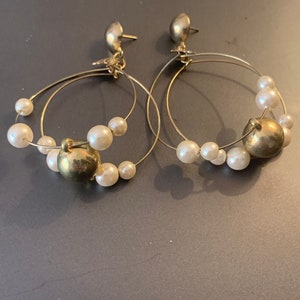May include: A pair of gold hoop earrings with a double hoop design and white pearl accents. The earrings have a gold ball charm on each hoop.