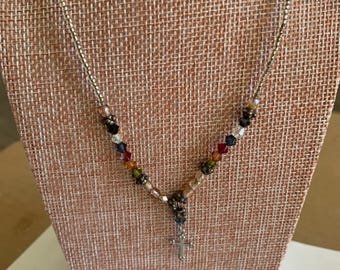 May include: A necklace featuring a silver-toned metal cross pendant suspended from a chain of small metallic beads. The lower section of the chain is accented with a series of multi-colored faceted crystal beads in shades of red, green, and blue.