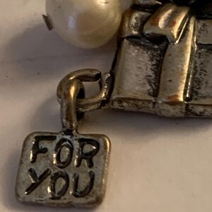 Puede incluir: Primer plano de un charm plateado con una perla y una peque&ntilde;a etiqueta rectangular. La etiqueta dice "FOR YOU". El charm tiene un dise&ntilde;o de caja de regalo.