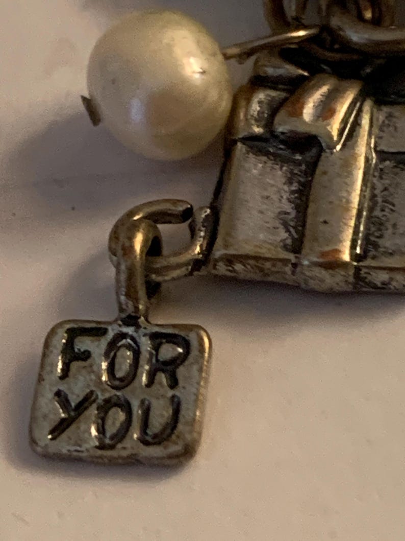 Puede incluir: Primer plano de un charm plateado con una perla y una peque&ntilde;a etiqueta rectangular. La etiqueta dice "FOR YOU". El charm tiene un dise&ntilde;o de caja de regalo.