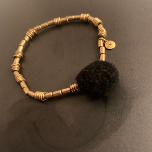May include: A gold bracelet with a black pom-pom charm. The bracelet is made of small, round beads that are connected by thin metal rings.