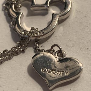 May include: Silver-toned necklace featuring a Mickey Mouse head pendant and a heart-shaped charm. The heart charm is engraved with the word "Disney". The necklace has a delicate chain.
