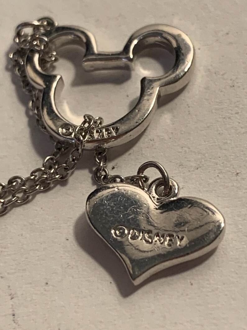 May include: Silver-toned necklace featuring a Mickey Mouse head pendant and a heart-shaped charm. The heart charm is engraved with the word "Disney". The necklace has a delicate chain.